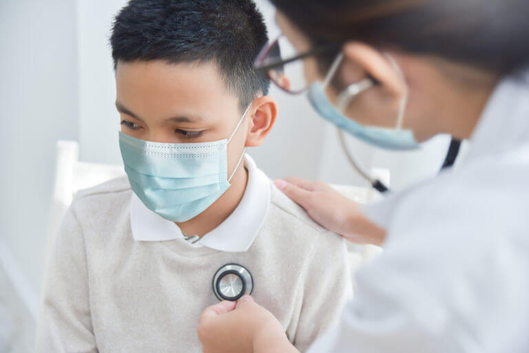 Doctor examining Asian boy wearing protective face mask with stethoscope